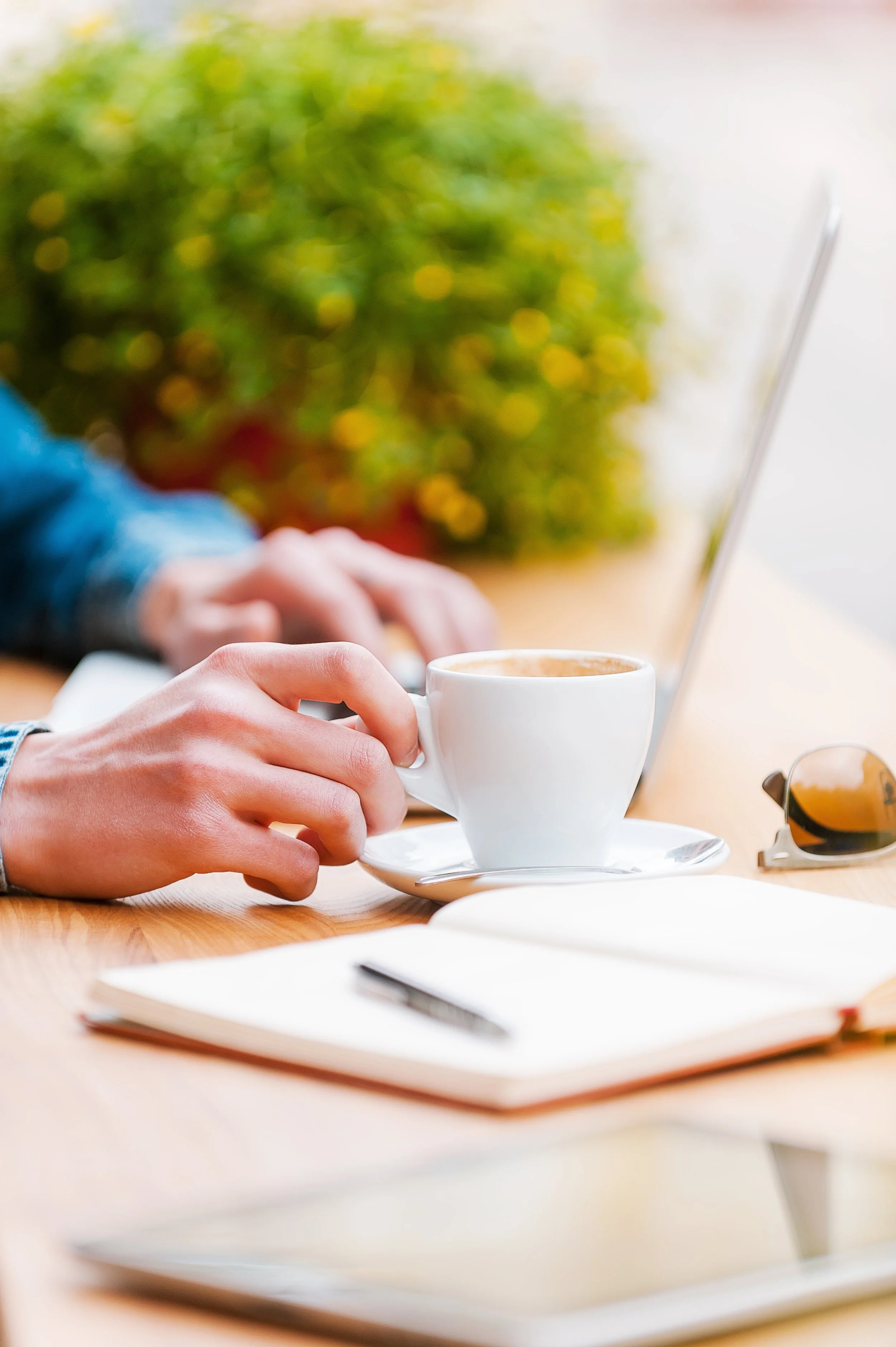 A person's hand holding a white coffee cup near a laptop on a wooden table, with a notebook and sunglasses nearby.
