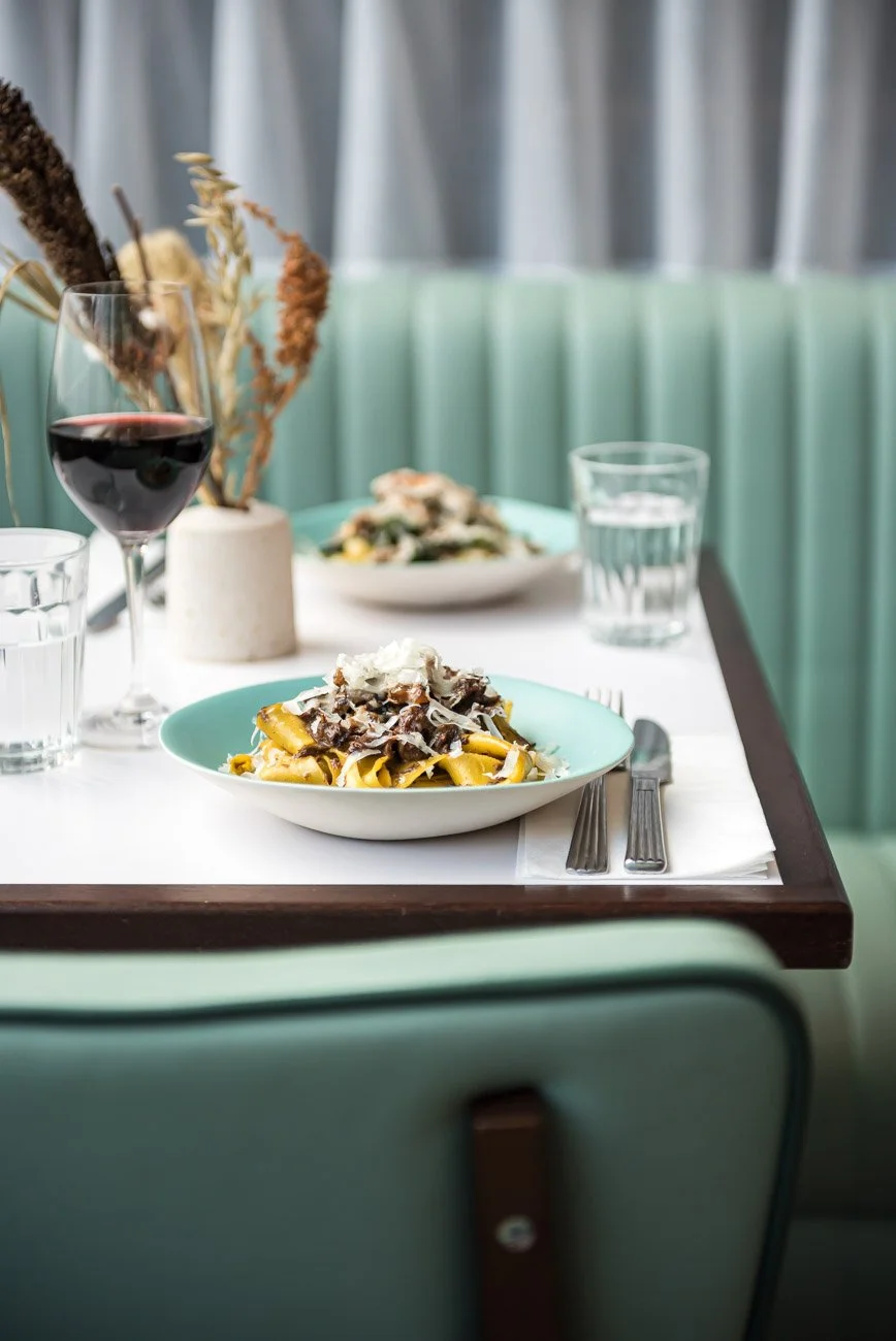 Pasta with shaved cheese and ground black pepper on mint green and white plates on a white table with silverware, glass of red wine, and glasses of water, set against a background of curtains and a teal booth.