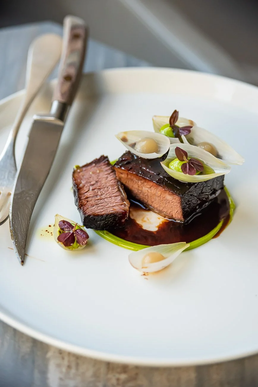 A plated dish of glazed beef with garnishes, including white and purple edible flowers and small green vegetables, on a white plate with cutlery nearby.