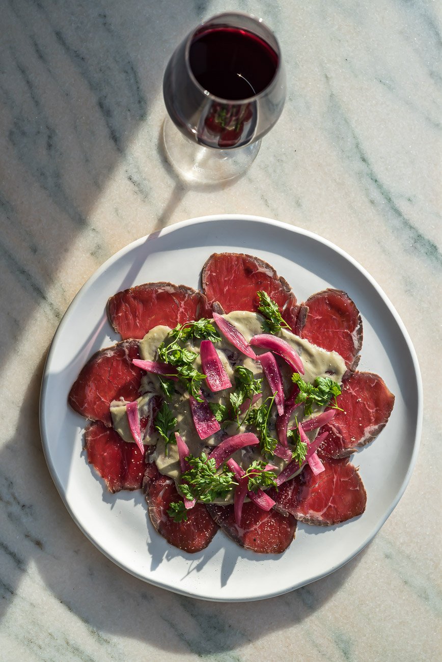 A white plate with raw beef carpaccio garnished with pink pickled onions and fresh herbs, served alongside a glass of red wine on a light-colored marble surface.