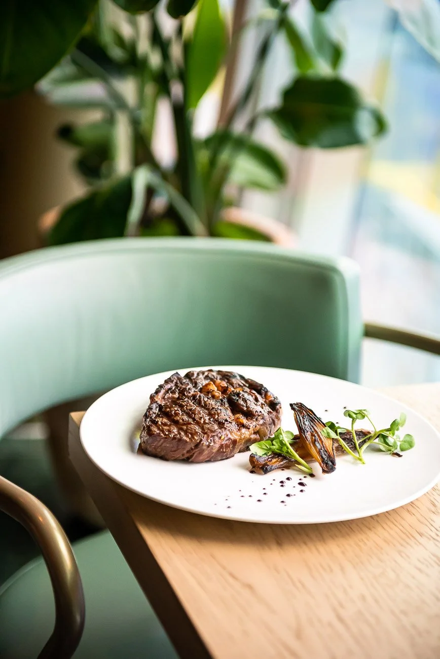 A plate with a cooked steak, grilled vegetables, microgreens, and black peppercorns on a wooden table, with a green chair and indoor plants in the background.