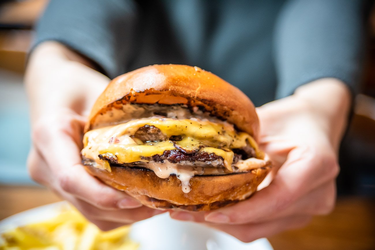A person holding a cheeseburger with two beef patties, melted cheese, and a toasted bun.