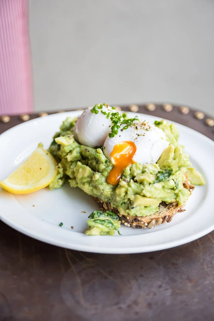 Sliced avocado toast topped with two poached eggs, garnished with chopped chives, with a lemon wedge on the side.