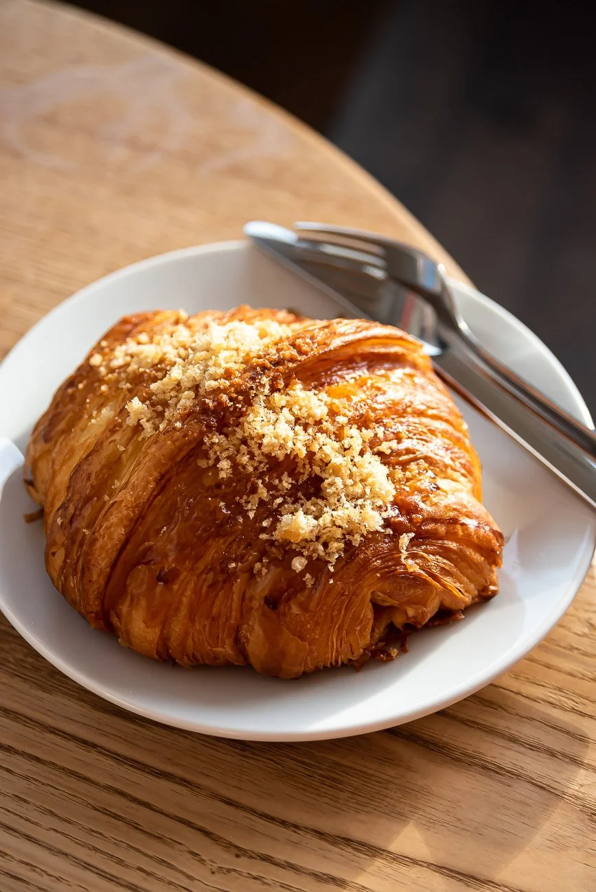 Close-up of a flaky, golden-brown croissant topped with crumbly streusel on a white plate, with a fork and knife placed beside.