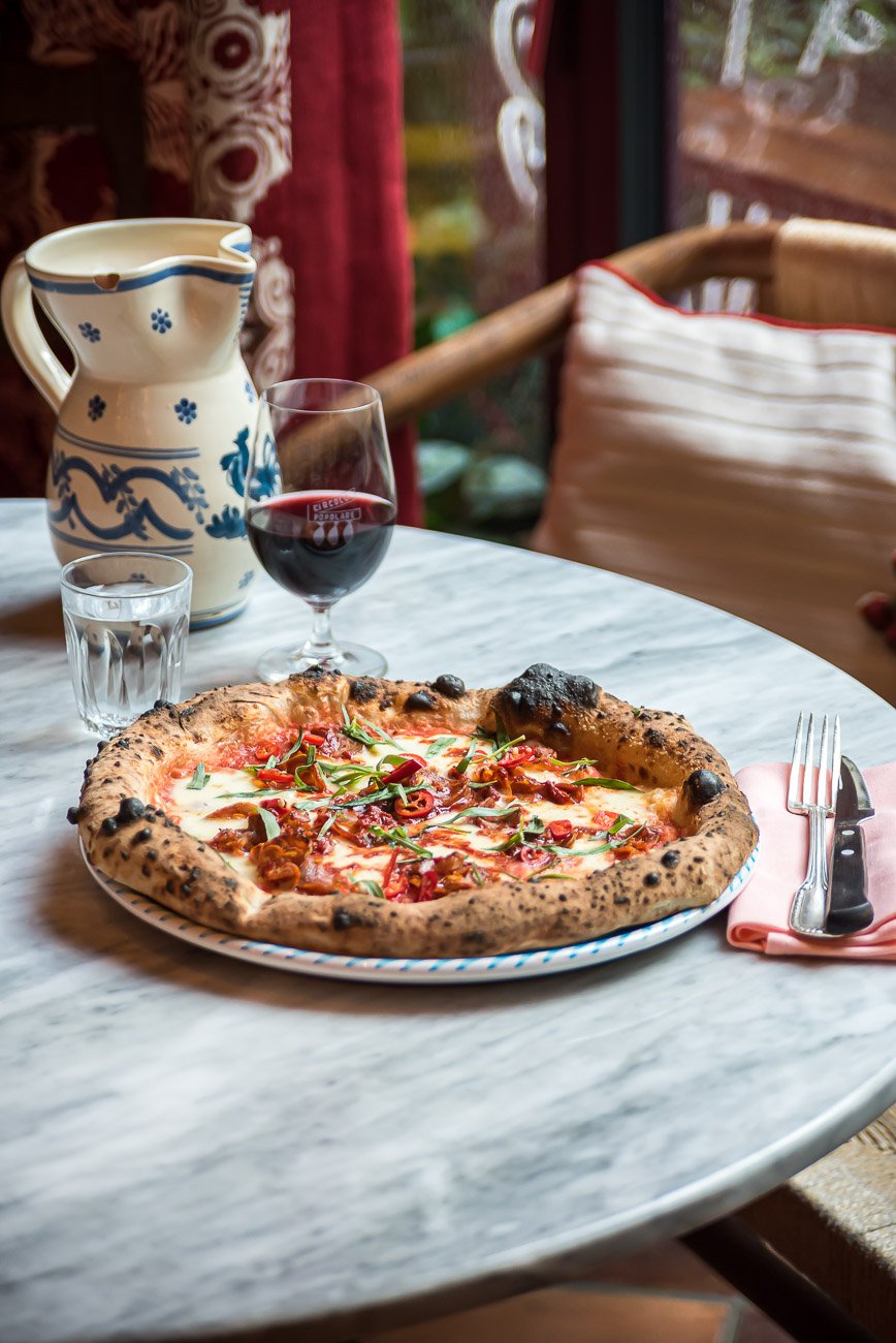 A marble table holding a freshly baked pizza with cheese, red peppers, and herbs, accompanied by a glass of red wine, a glass of water, a ceramic pitcher with blue patterns, and silverware on a pink napkin.