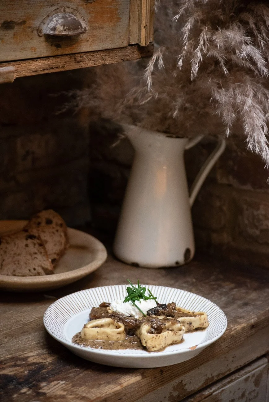 A plate of pasta with sauce and herbs, served on a rustic wooden table next to a vase with dried pampas grass, with a plate of sliced bread nearby.