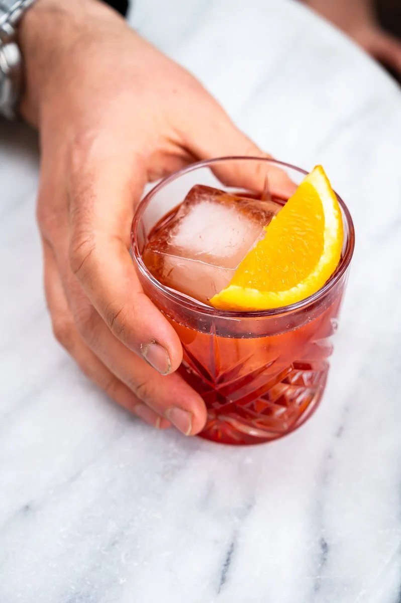 A person holding a glass of pink-colored cocktail with a large ice cube and a slice of orange garnish on a white marble surface.