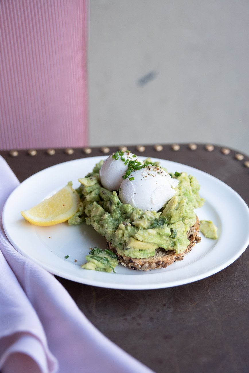 Poached eggs on a bed of mashed avocado on toast with a lemon wedge, garnished with chives, served on a white plate.