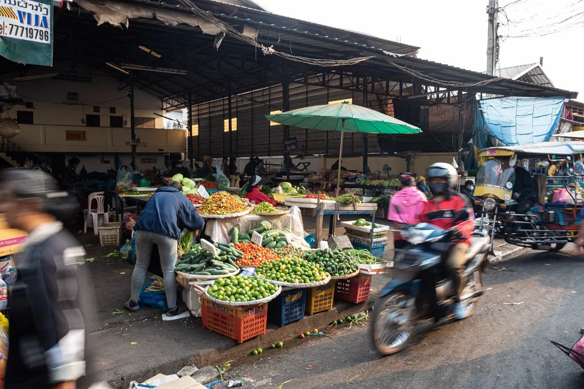Street market selling various fresh vegetables and fruits, with vendors and customers, covered by a large umbrella, and moving vehicles including motorcycles and tuk-tuks.