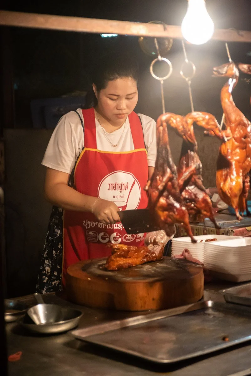 Woman slicing roasted chicken at a food stall, with cooked chickens hanging above.