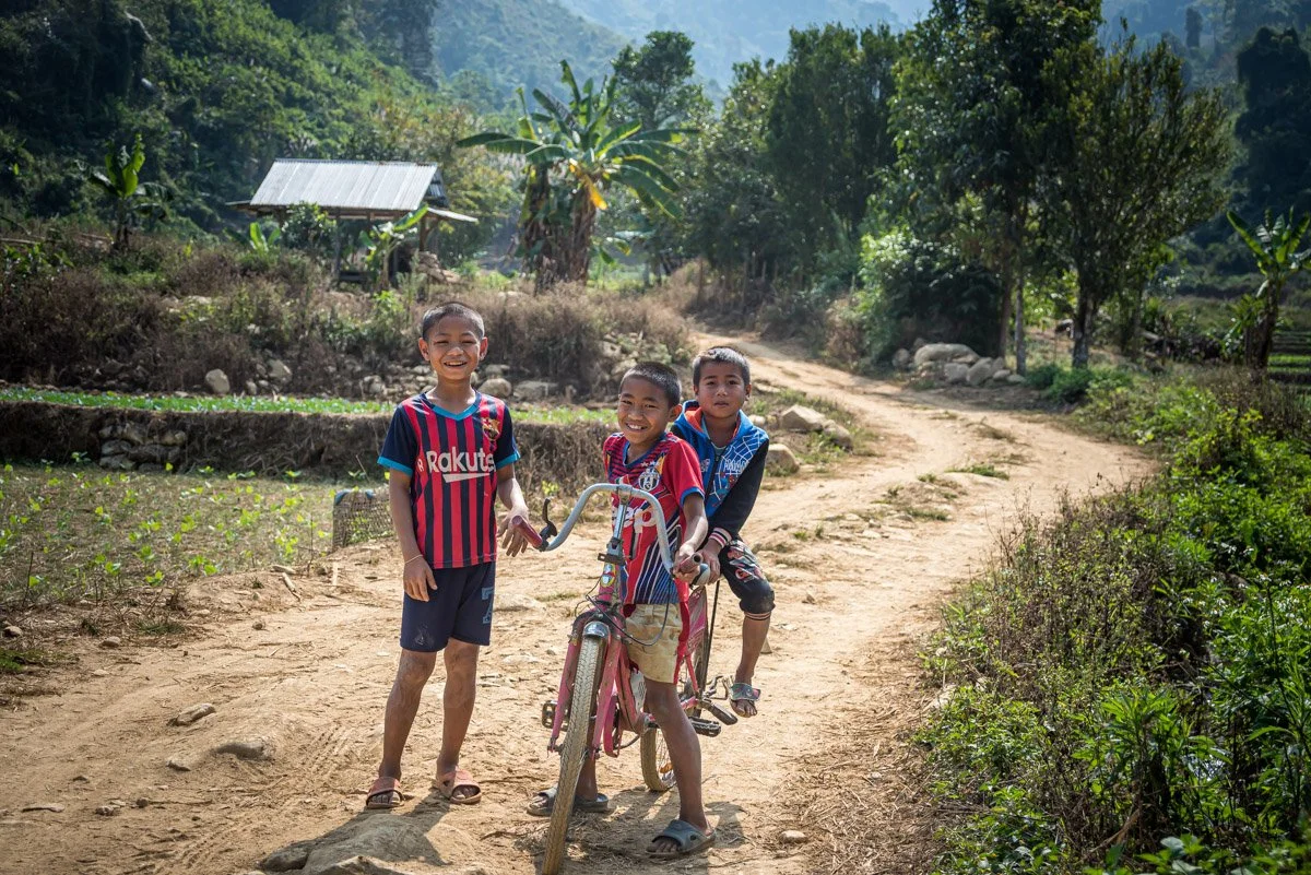 Three young boys smiling and playing on a dirt path in a rural area with trees and a small wooden structure in the background.
