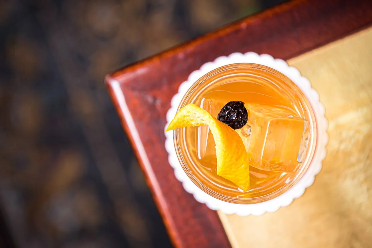 A glass of iced orange drink with a lemon wedge and a black berry, placed on a white doily on top of a wooden table.