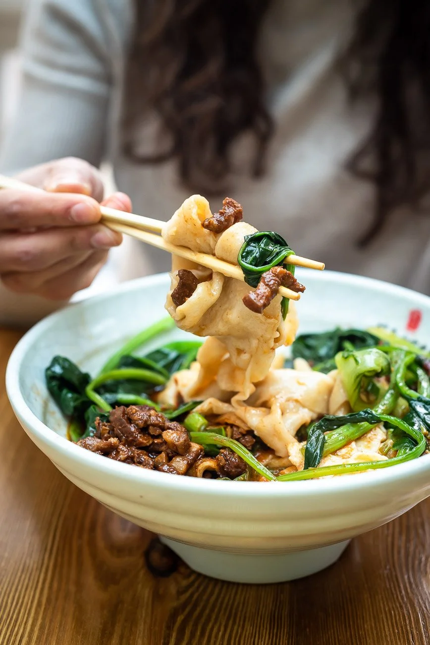 A person holding chopsticks with a large piece of dumpling, some beef, and green leafy vegetables above a bowl of similar ingredients on a wooden table.