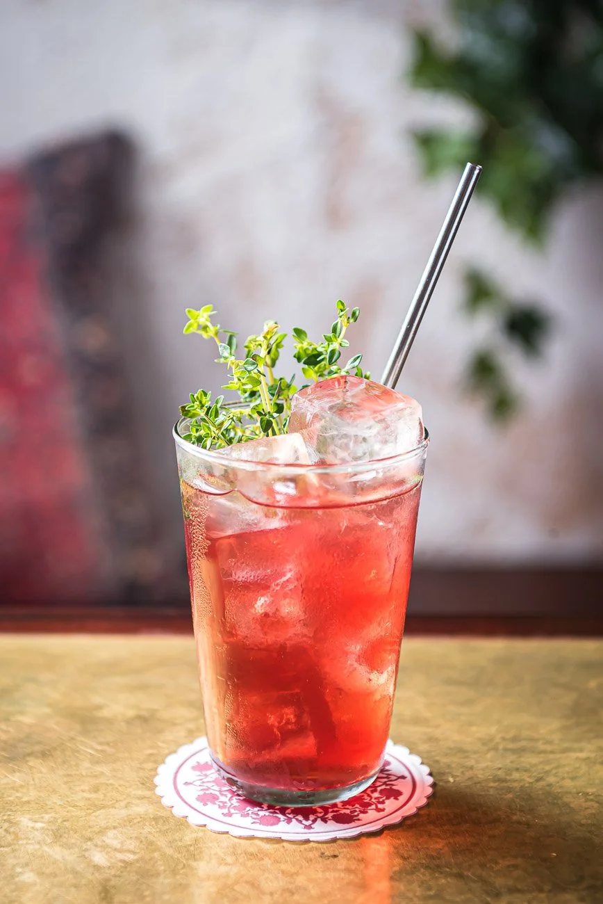 A tall glass of red-colored iced beverage garnished with greens and a metal straw, placed on a white coaster with a floral pattern on a wooden surface.