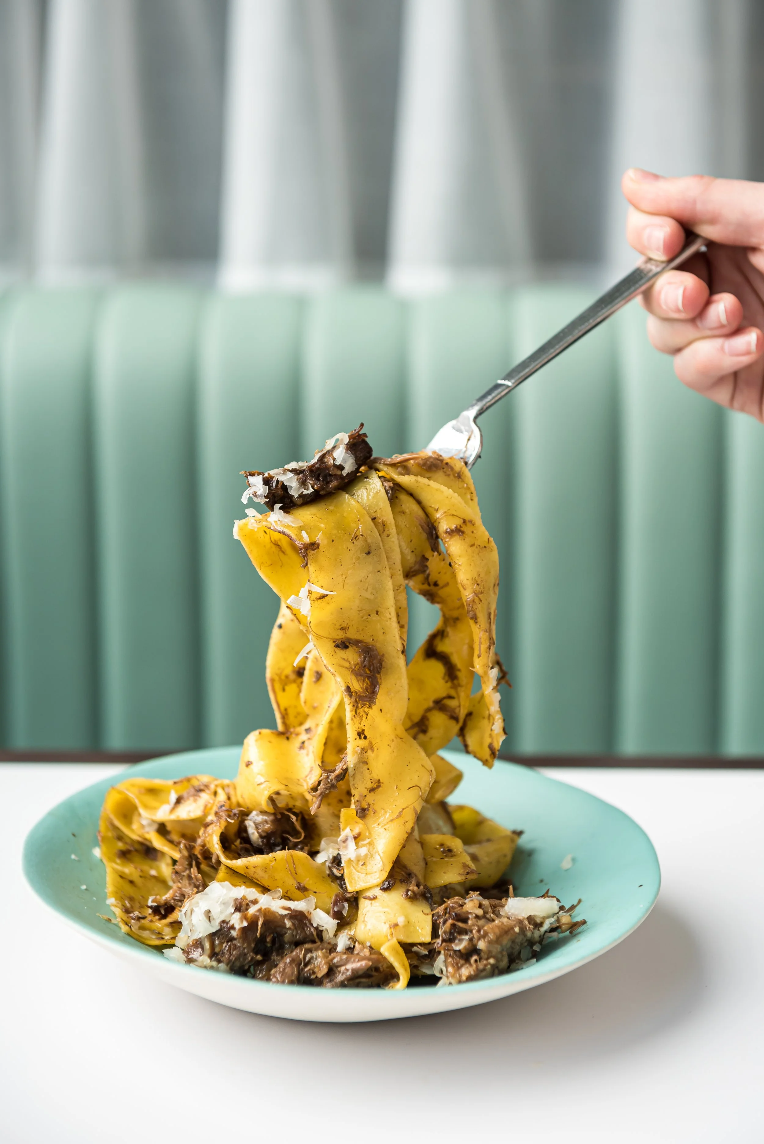 A person is lifting a serving of banana foster pasta with a fork onto a turquoise plate. The pasta is yellow with dark chocolate and shredded coconut toppings, set against a blurred background of a metallic wall with vertical ridges.