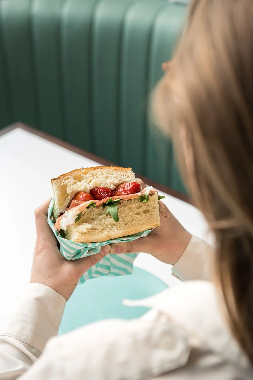 Person holding a large sandwich with cherry tomatoes, arugula, and sliced meat, seated at a white table with green booth seating in the background.