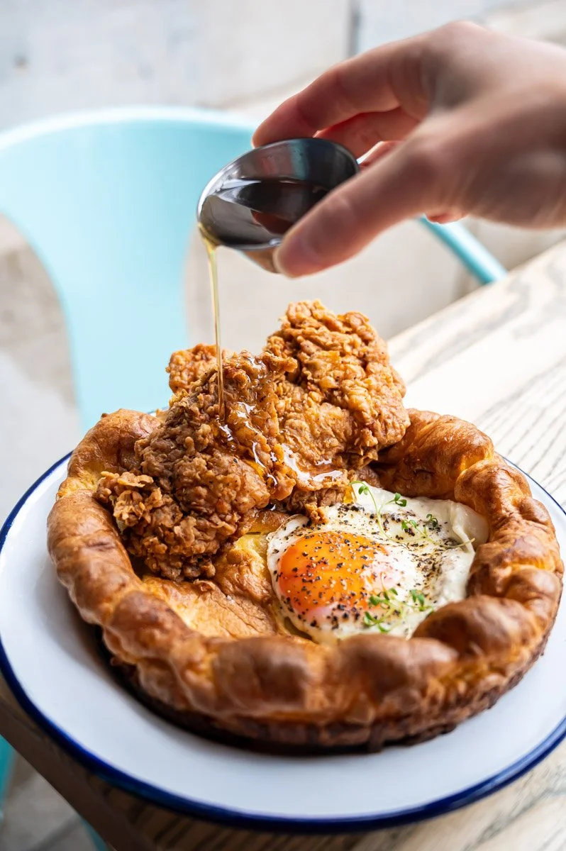 Fried chicken with a fried egg in a biscuit bowl, topped with syrup being poured from a small pitcher.