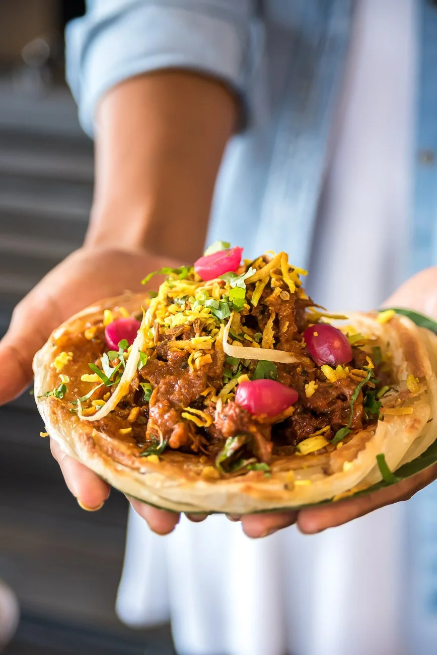 A person holding a traditional Mexican birria taco filled with shredded meat, topped with chopped onions, cilantro, shredded cheese, and garnished with Pickled red onions and radishes.