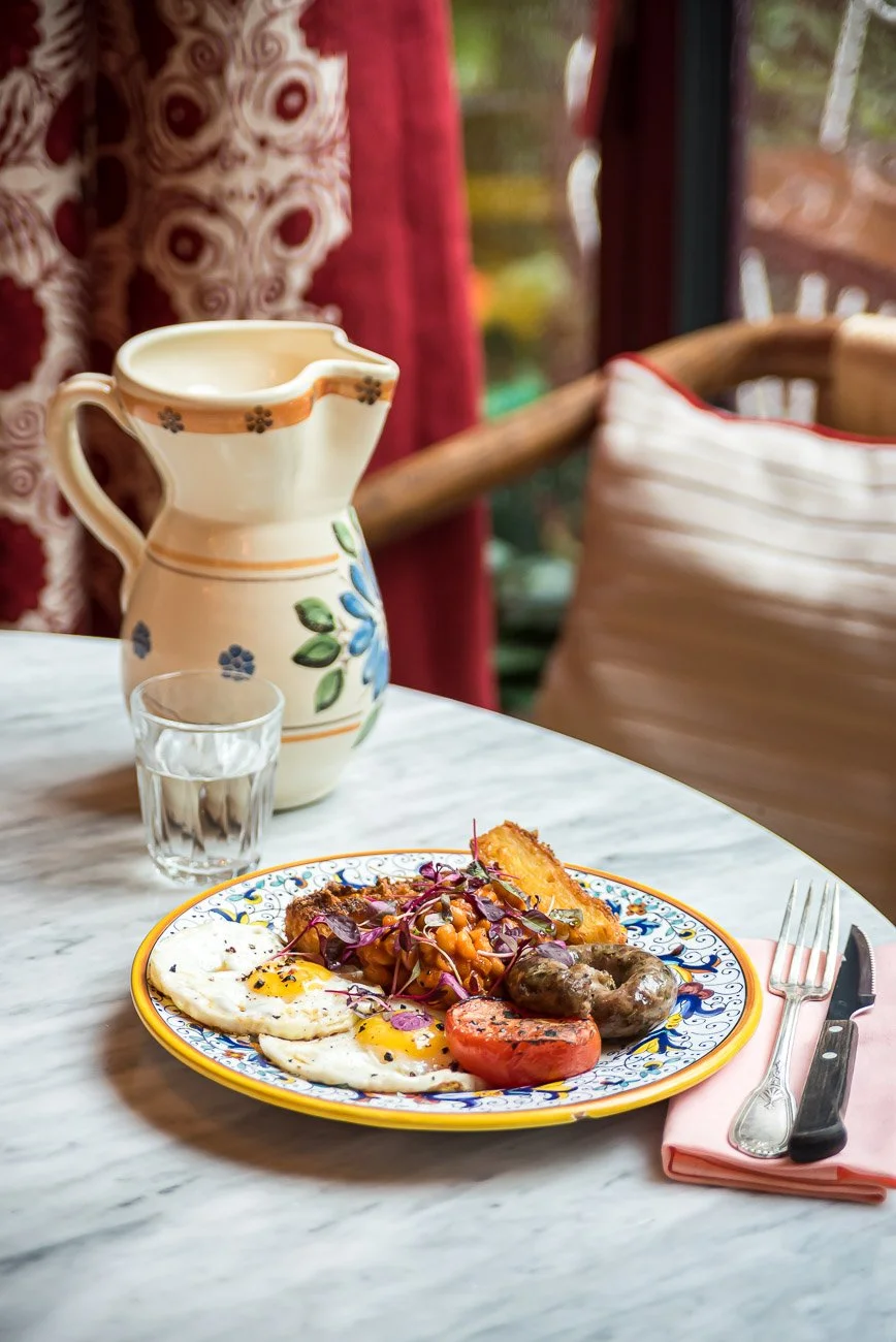 A breakfast plate with sunny-side-up eggs, grilled tomato, sausage, hash browns, and sautéed vegetables on a colorful plate. A glass of water, a ceramic pitcher, and a fork and knife on a pink napkin are on a white marble table. Red curtains and a wi