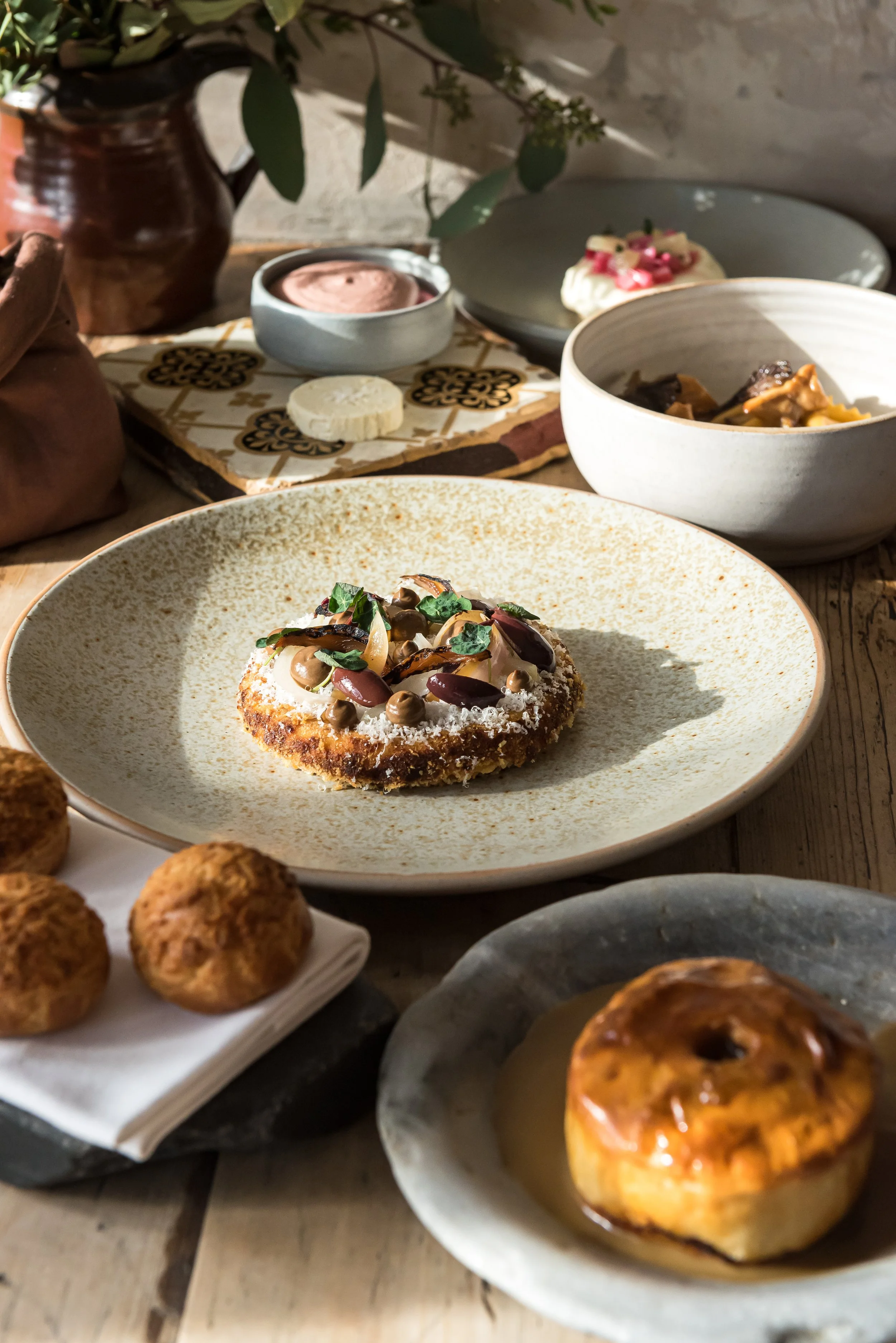 An assortment of desserts including a decorated donut topped with chocolate and herbs, a pastry with caramelized topping, and small round cookies or balls on a white plate, arranged on a wooden table.