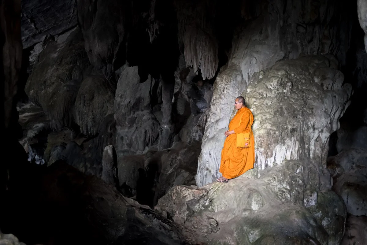 A Buddhist monk dressed in orange robes standing inside a dark cave on a large rock formation.