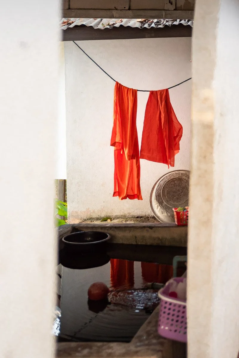 Orange and red cloths hanging on a clothesline outdoors, with a washing area below, reflecting the hanging clothes in water, and a small pink basket in the foreground.