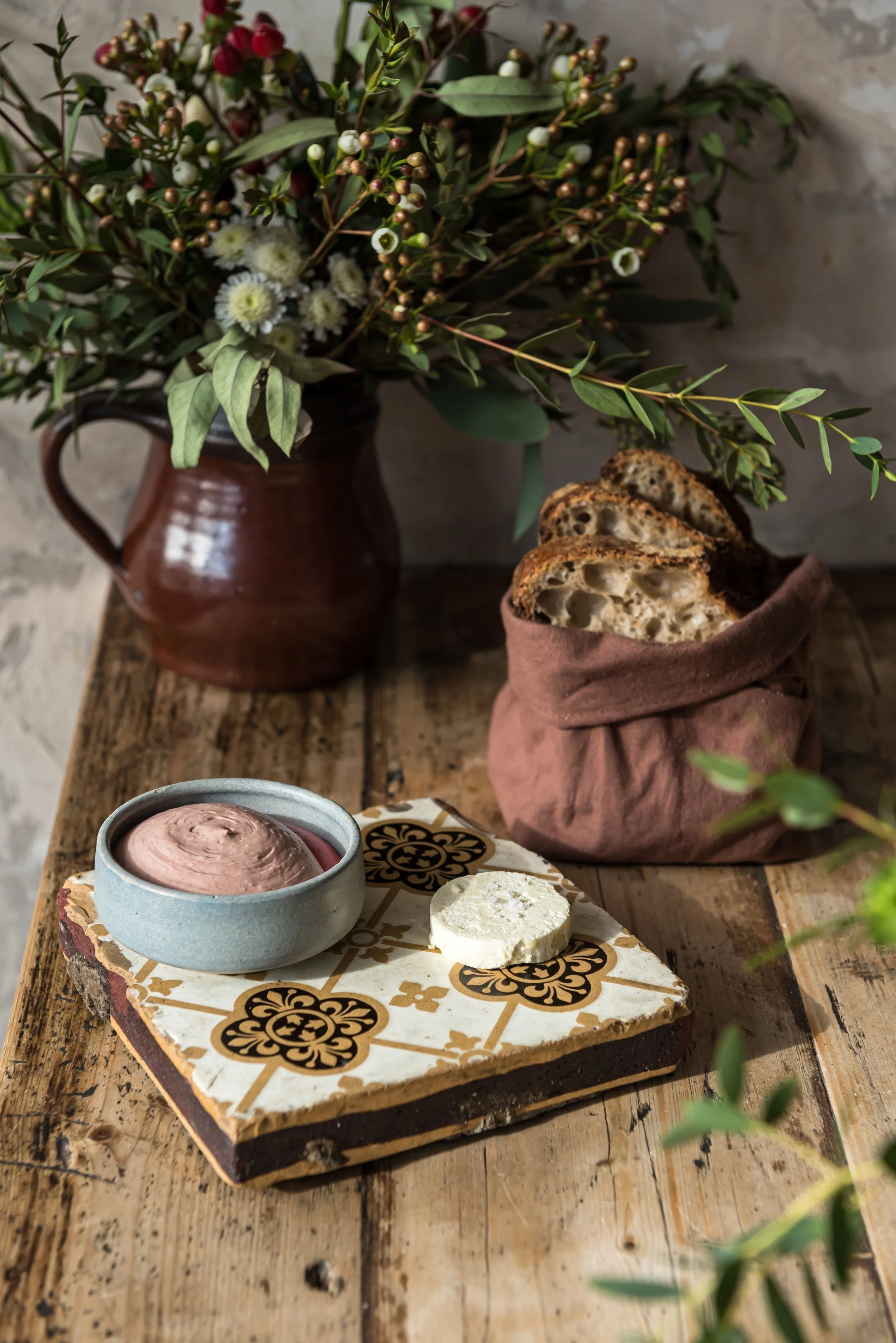 A rustic wooden table features a small blue ceramic bowl filled with chocolate mousse, a white cheese, and a patterned tile serving platter. In the background, an earthenware pitcher contains a lush bouquet of greenery and white flowers, with a wrapp