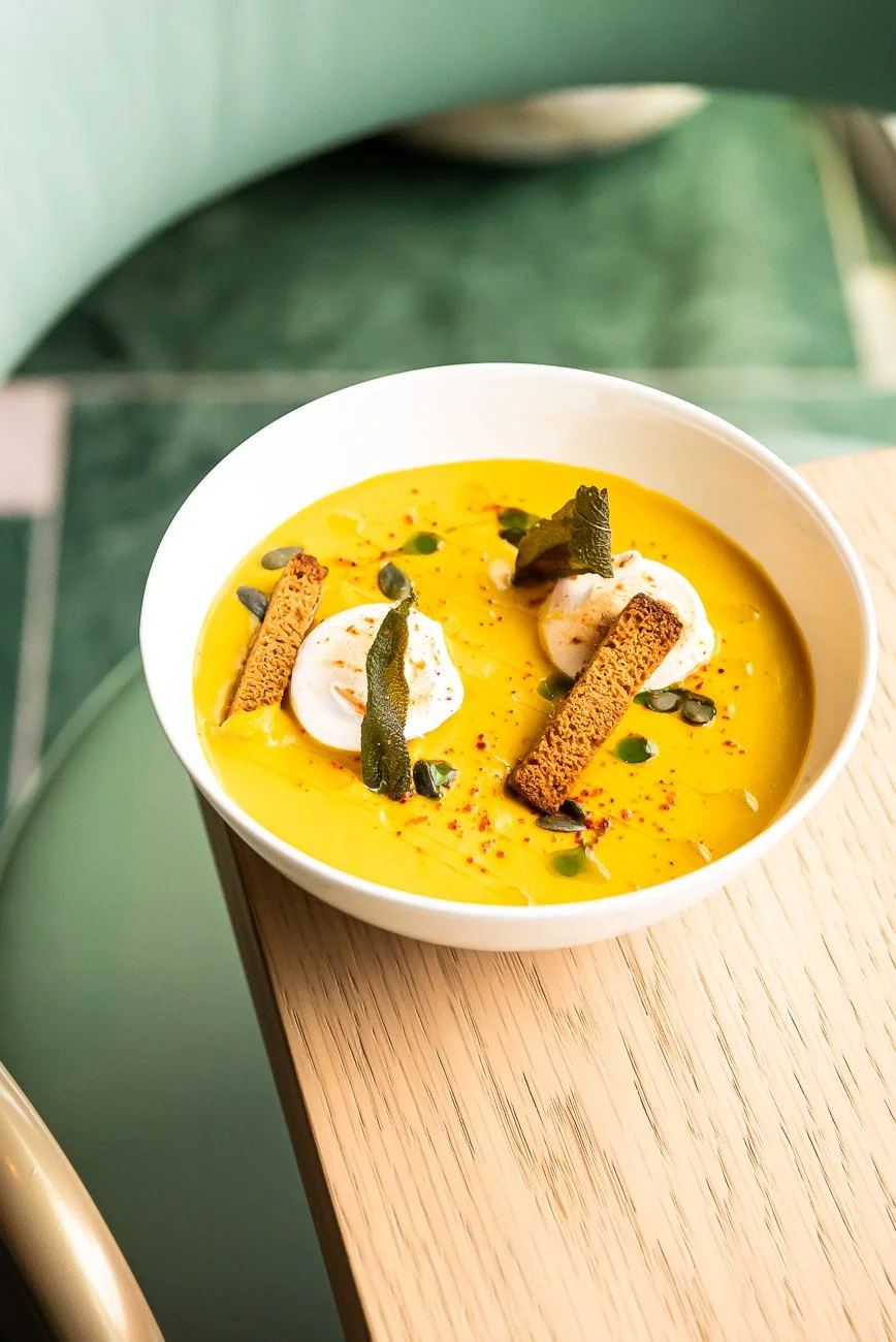 A bowl of yellow soup garnished with dollops of cream, toasted bread sticks, pumpkin seeds, green herb leaves, and red chili flakes.