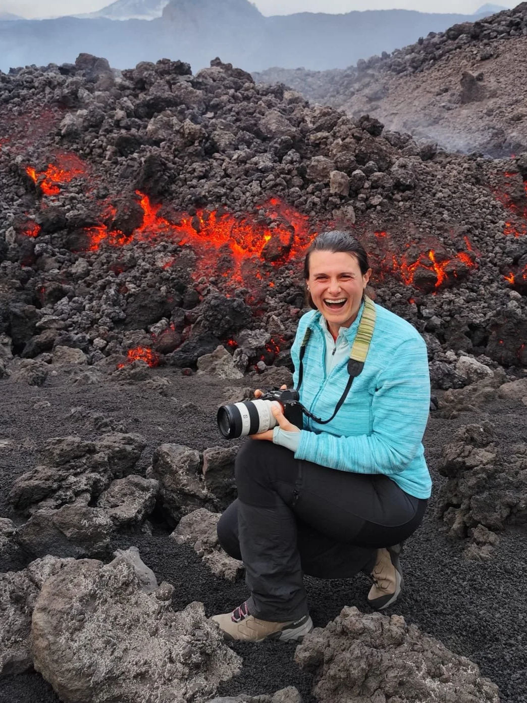 A woman in outdoor gear smiling and holding a camera, kneeling on volcanic rocks with glowing lava and smoke in the background.
