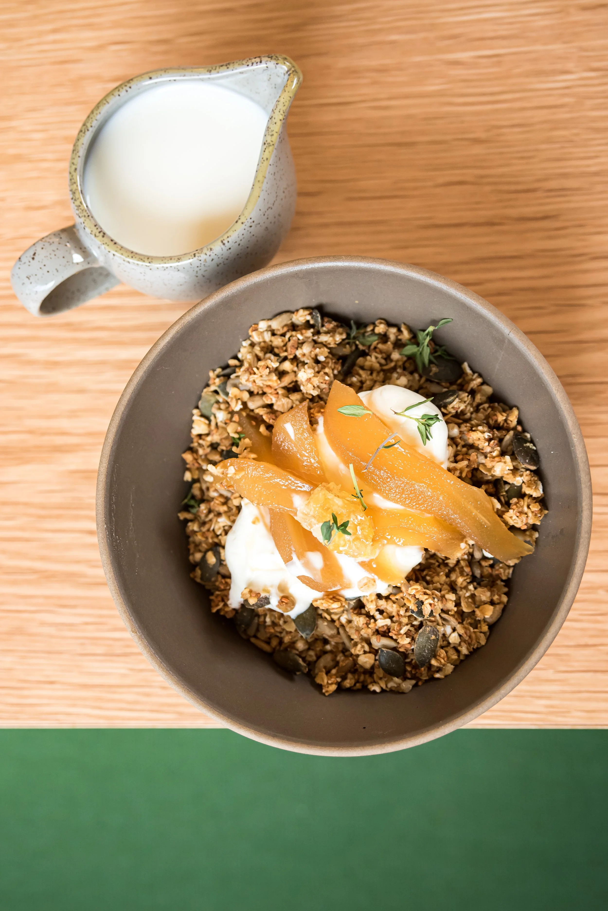 A bowl of granola with yogurt and fruit topping, and a pitcher of milk, on a wooden table.
