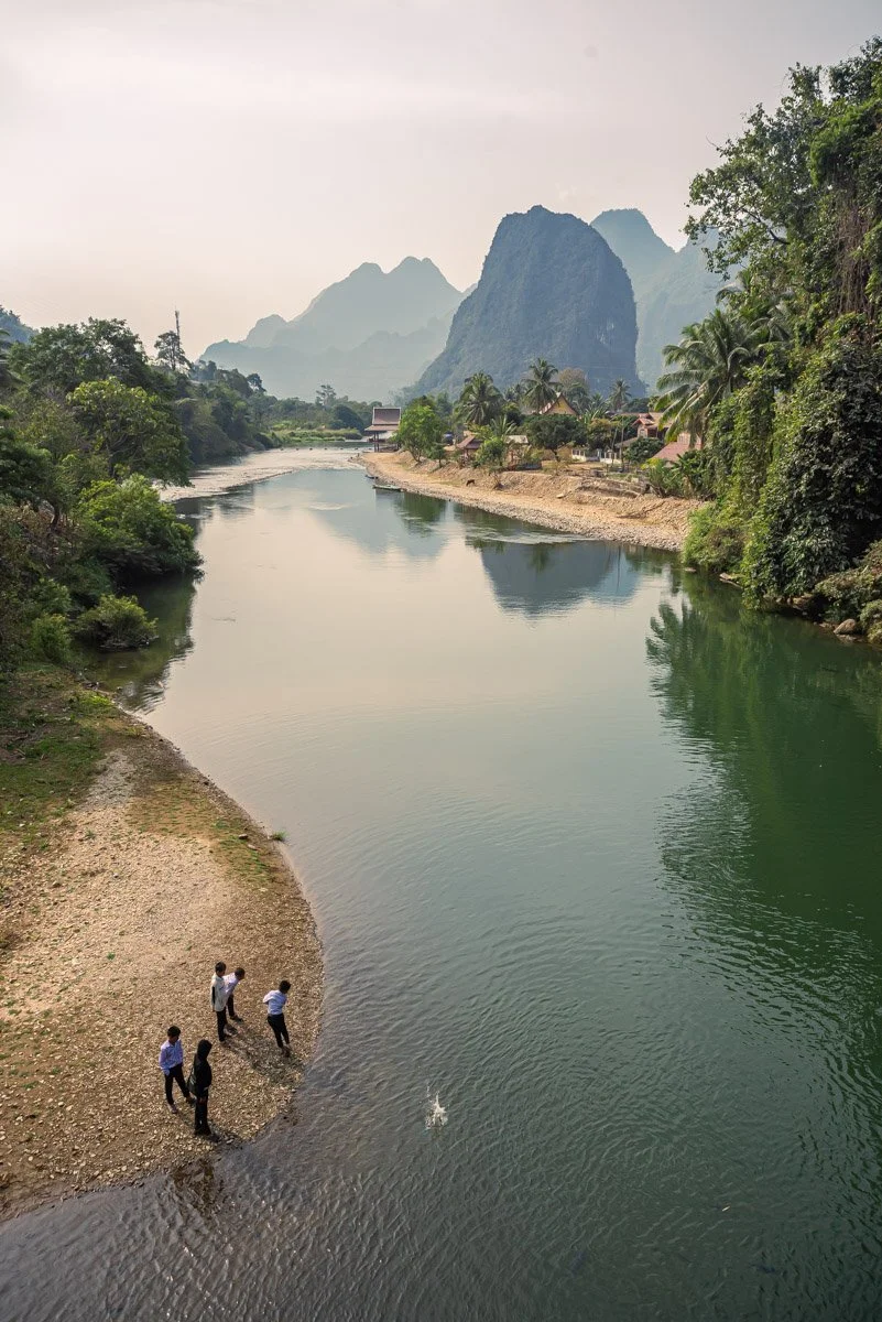 Four people standing on a pebbled riverbank with lush green trees and mountains in the background.