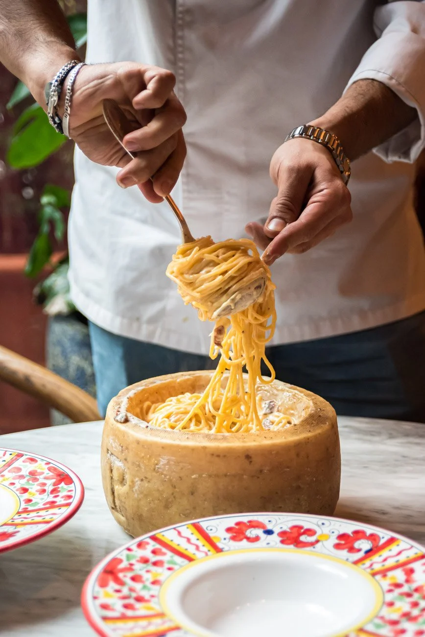 A person serving spaghetti from a hollowed-out cheese wheel onto decorative plates.