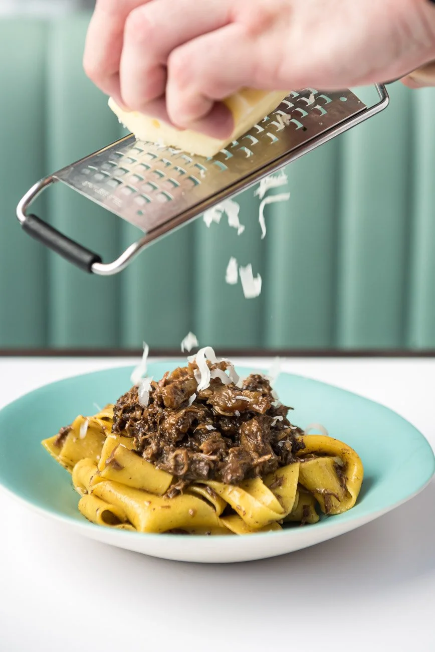 Hand grating cheese over a plate of pasta with a dark meat sauce