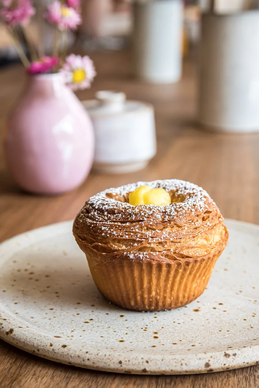A croissant-style pastry with powdered sugar and a dollop of butter on top, placed on a speckled white plate, with a blurred pink vase with flowers and white jars in the background.