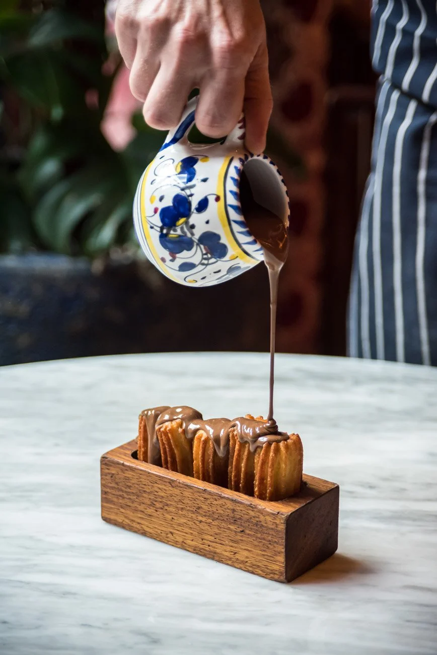 A person pouring chocolate sauce from a decorative cup over churros arranged in a wooden tray on a white marble surface.