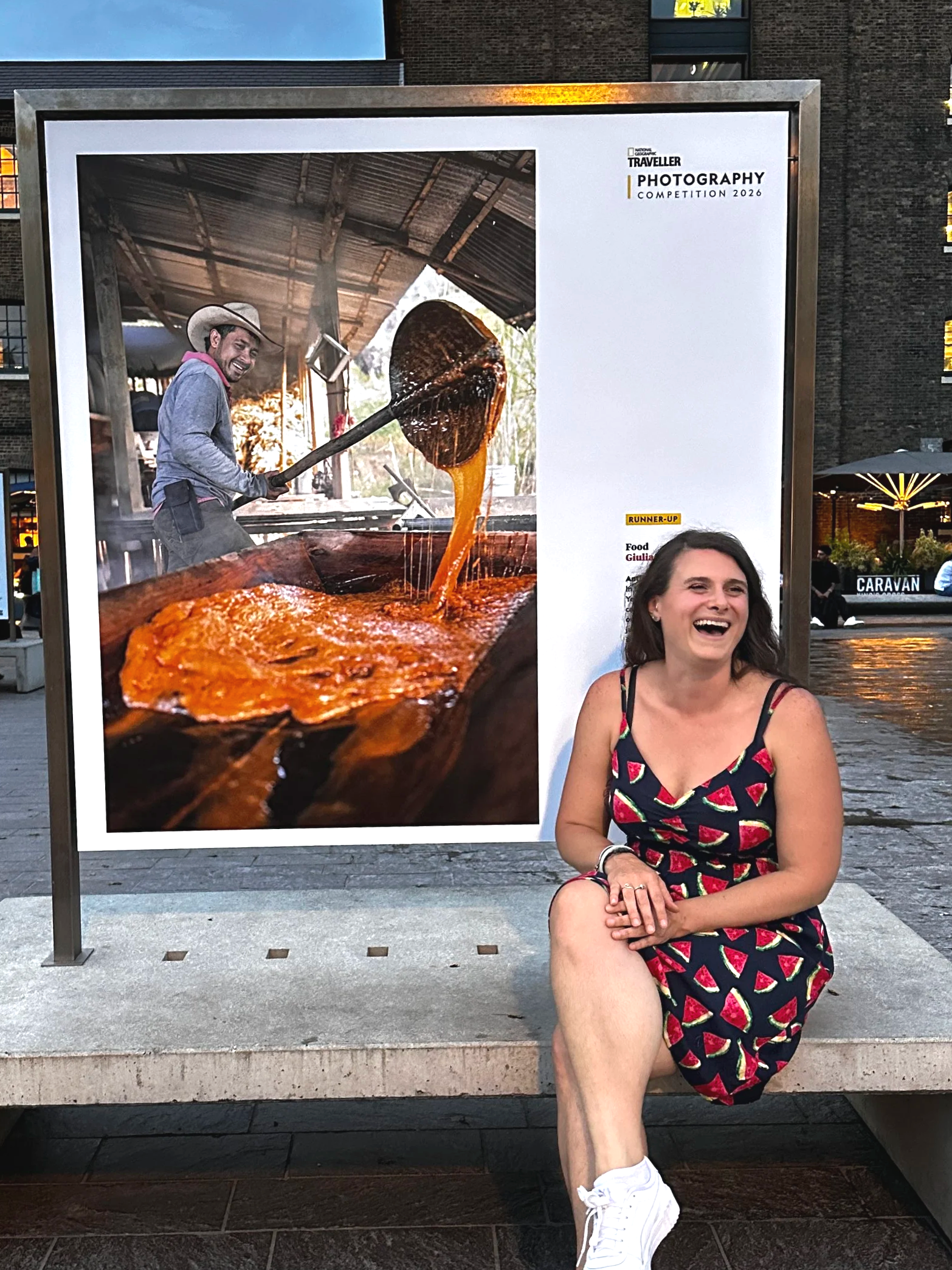A woman sitting on a bench with a large photographic poster behind her. The poster features a man cooking, holding a large spatula in a rustic open-air kitchen with a large pot of food.