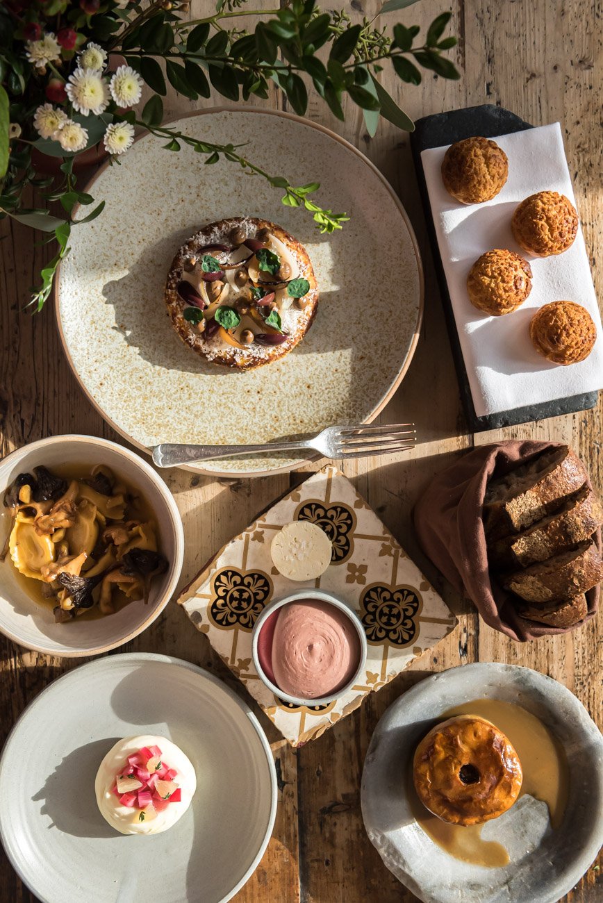 A variety of desserts on a rustic wooden table, including a pastry with chocolate and sprinkles, three coconut macaroons, a loaf of sliced bread, a bowl of mushroom pasta, a small bowl of pink mousse, a cupcake with pink flower decorations, and a gla