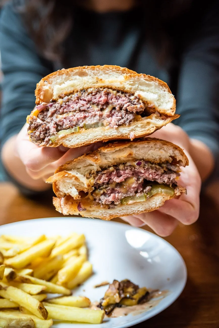 Person holding a cheeseburger with a beef patty, cheese, pickles, and sauce, cut in half, with fries on a white plate below.