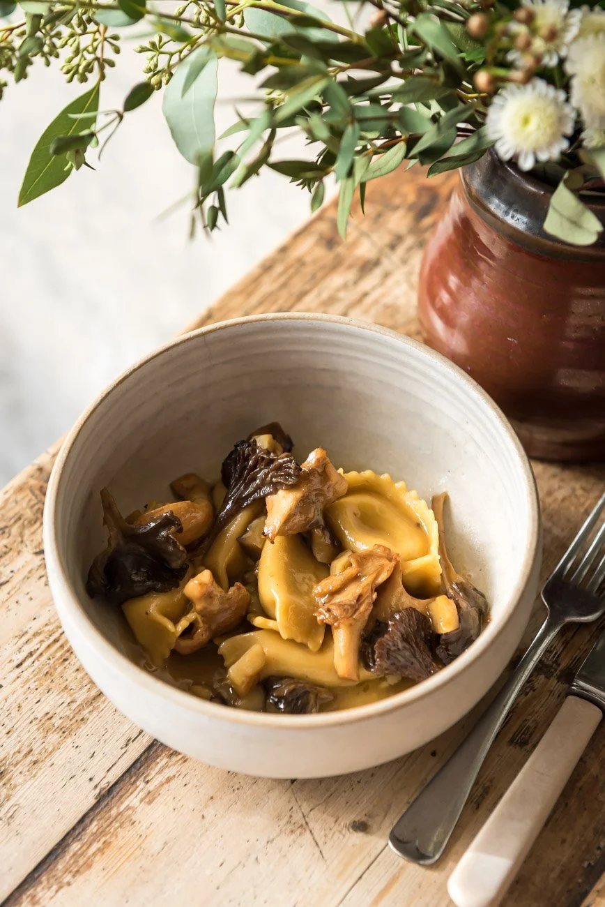 A bowl of cooked pasta with mushrooms on a rustic wooden table, next to a silver fork and a red vase with white flowers and green foliage.