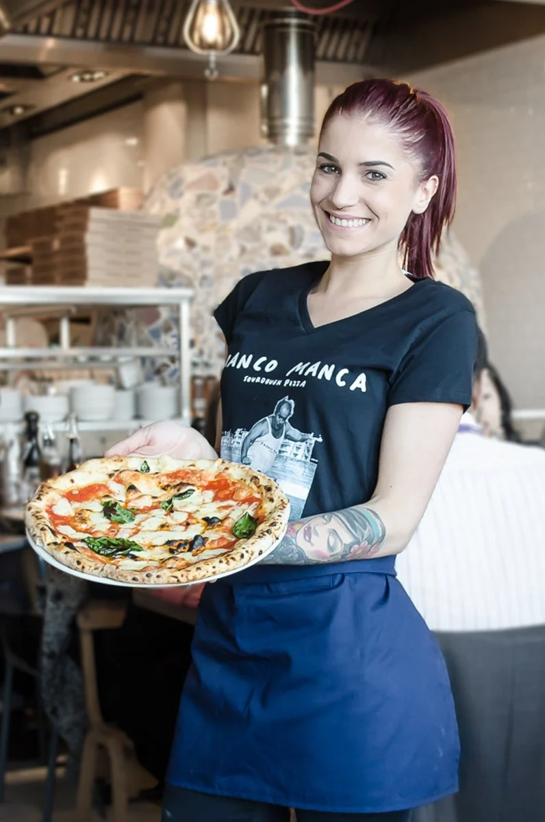 A woman with red hair, wearing a black T-shirt and blue apron, holding a pizza in a restaurant setting.