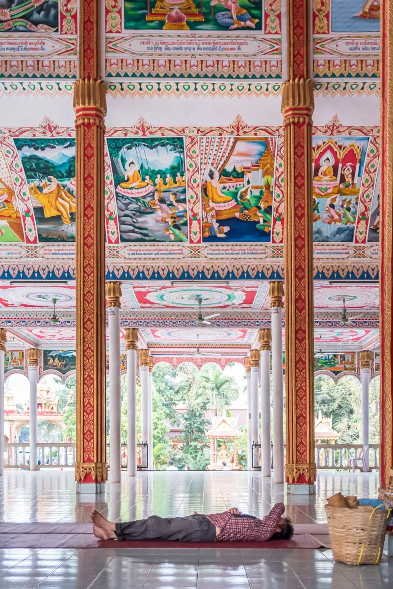 A person lies on their back on a yoga mat inside a colorful, ornate temple with intricate paintings on the ceiling and large open windows showing greenery outside.