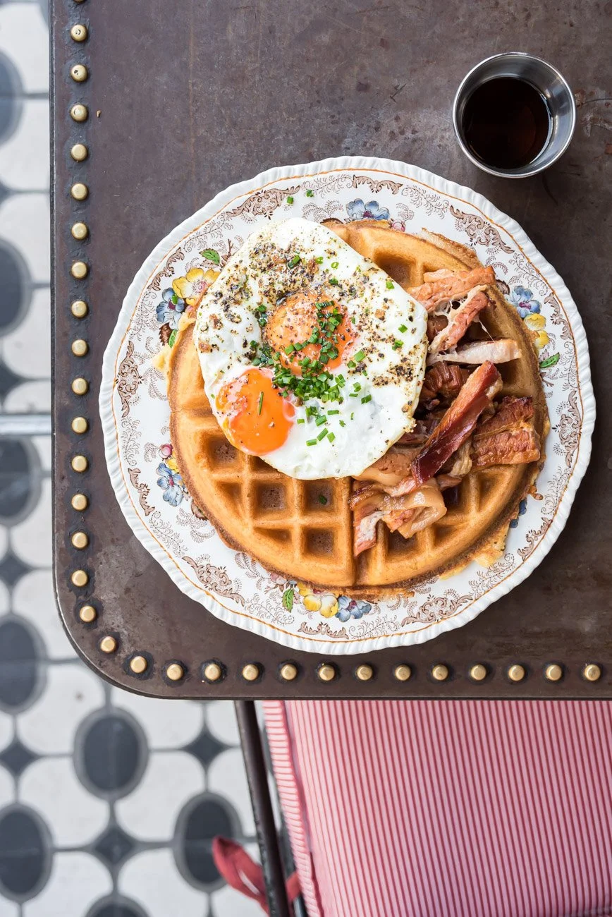 A plate of Belgian waffles topped with sunny-side-up eggs, chopped green onions, and black pepper, served alongside crispy bacon and a side of smoked salmon. There is a small cup of black coffee on the side, on a dark brown table with a decorative ed