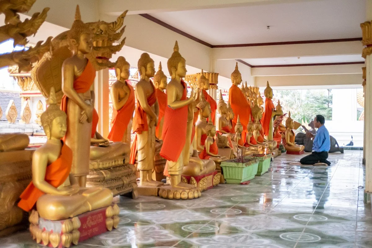 A person kneeling and praying in front of a row of golden Buddha statues inside a temple, with some statues wearing orange robes and others bare, arranged on raised platforms.