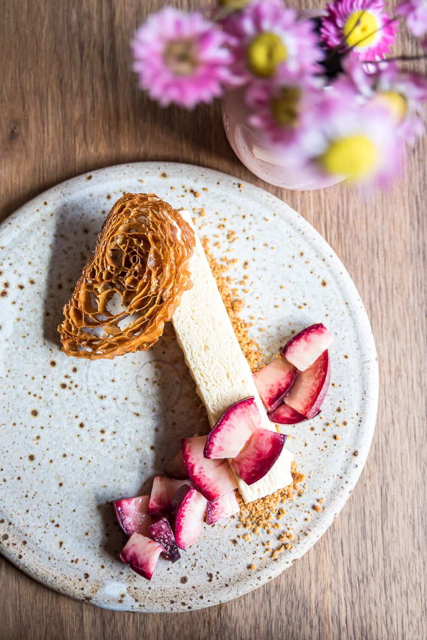 A dessert plate with a slice of cheesecake, topped with caramel honeycomb, served with sliced radishes, on a speckled white plate with a small pot of pink and purple flowers in the background.