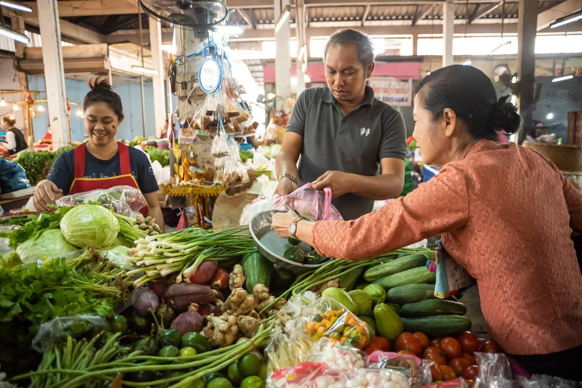 People buying fresh vegetables at a market stall. The stall has lettuce, cucumbers, tomatoes, ginger, and green onions on display.