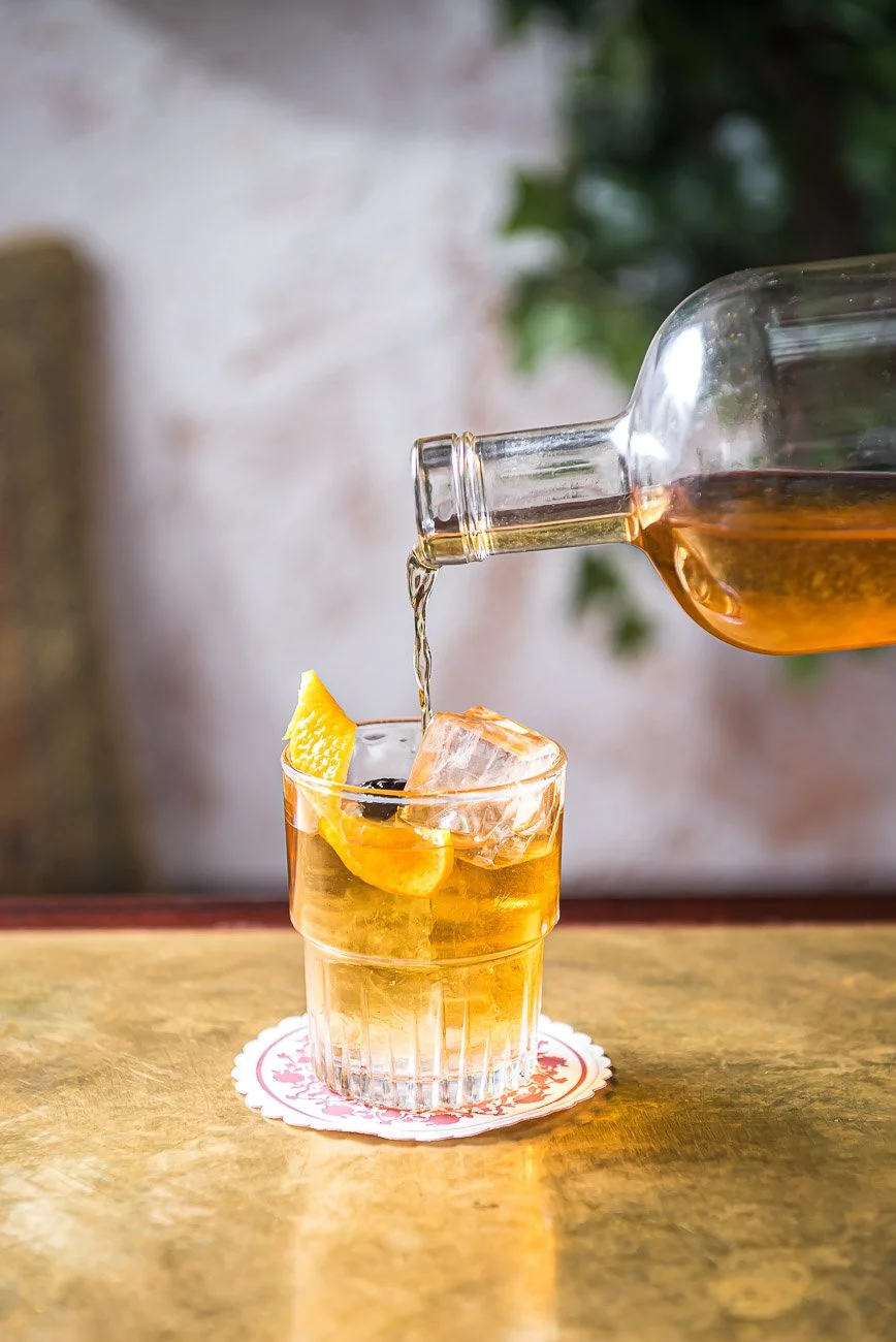 A glass of whiskey with ice and an orange twist, being poured from a glass bottle onto a wooden table with a decorative coaster.