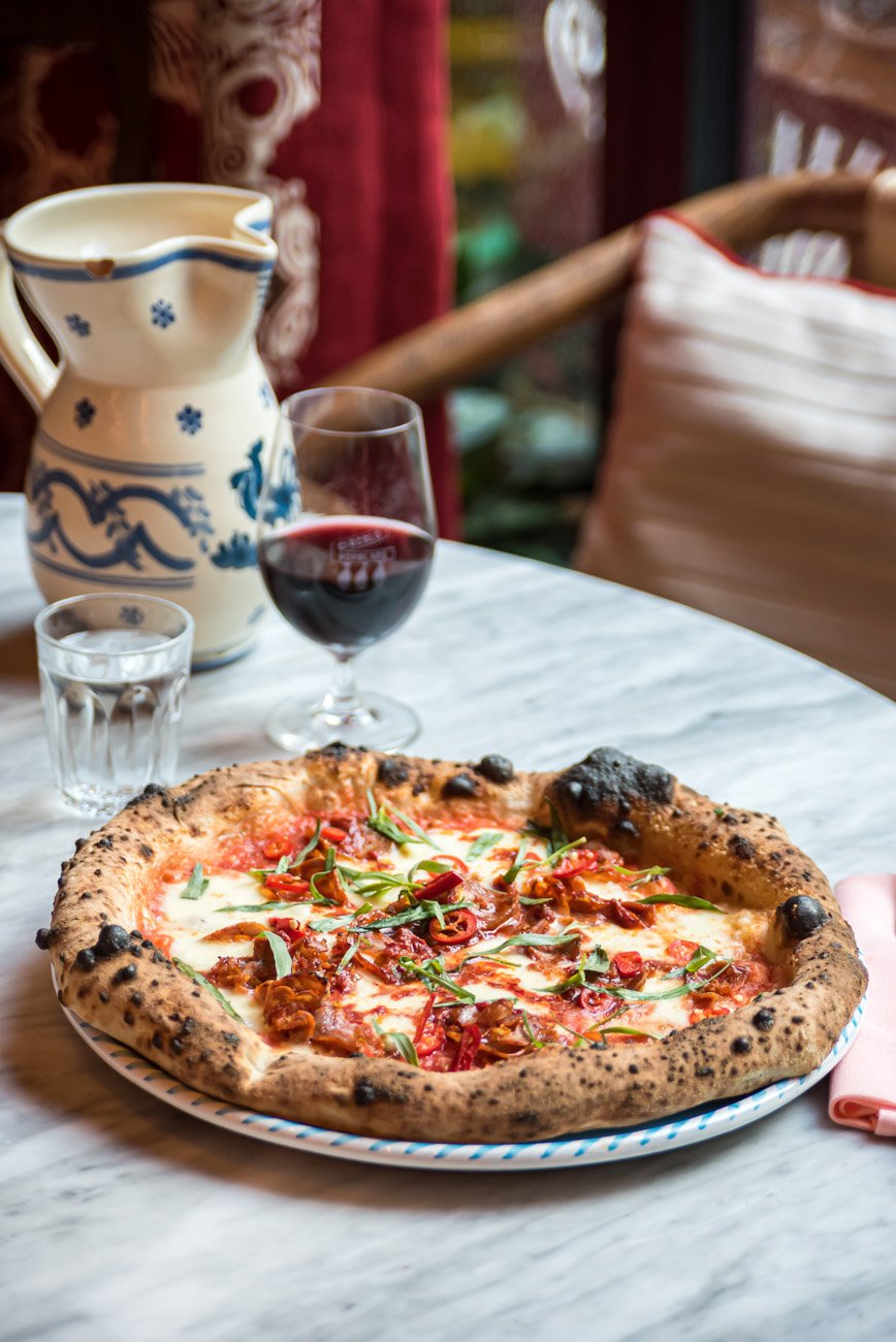 A pizza with cheese, red chili peppers, and fresh basil on a plate on a marble table. In the background, there's a glass of red wine, a water glass, a ceramic pitcher, and a cozy restaurant setting with colorful cushions and curtains.