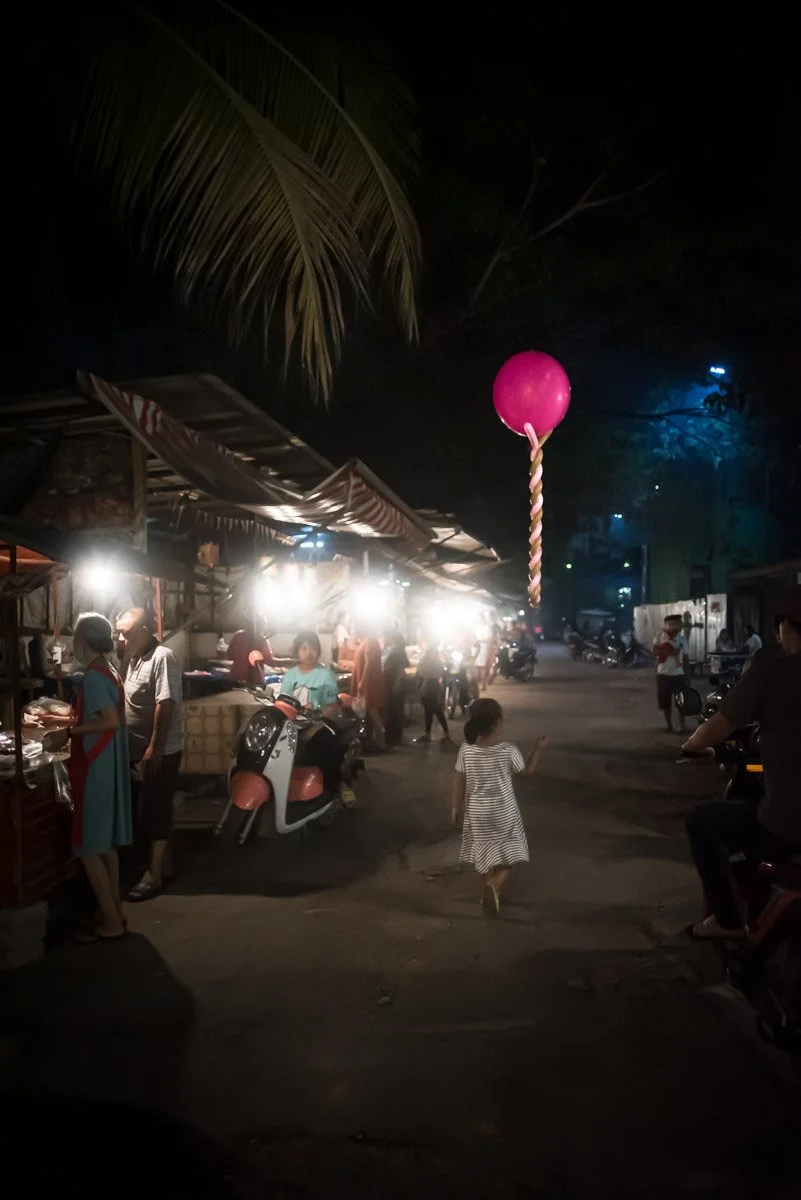 Night market scene with street vendors, people, and a child running holding a stick with a pink balloon, illuminated by bright lights, and a large palm leaf overhead.