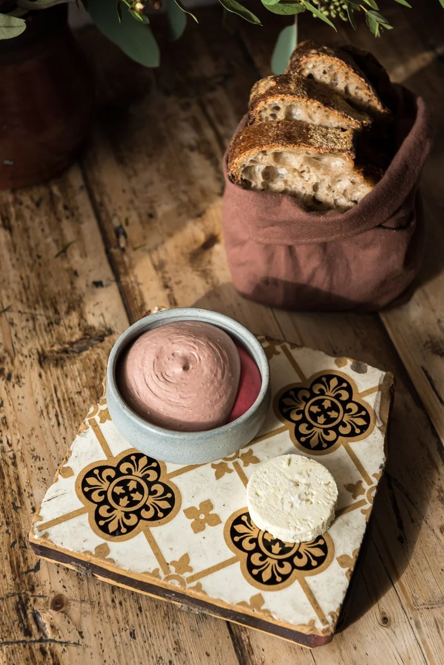 A rustic wooden table holding a small bowl of pinkish-brown mousse, a round white snack, and patterned paper with decorative design. Behind it, there is a brown paper bag with slices of toasted bread with visible holes and a crunchy crust, placed in 