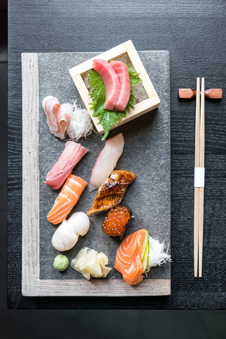 Assorted pieces of sushi arranged on a black and gray serving board, with chopsticks to the right.
